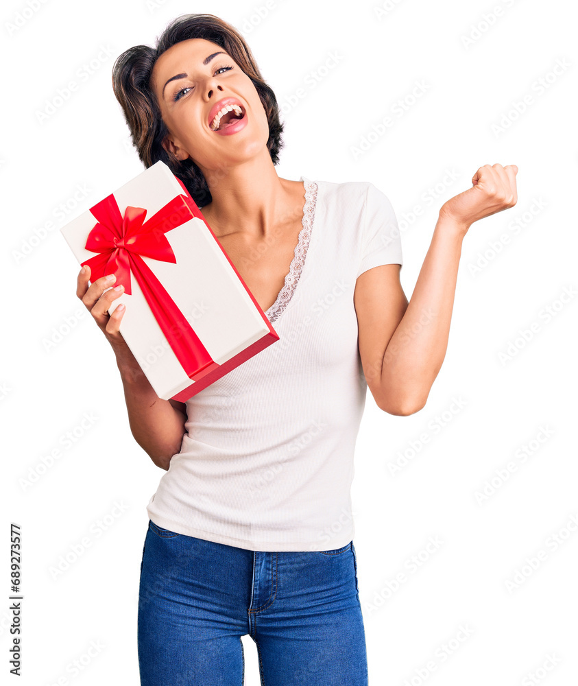 Young beautiful woman holding gift screaming proud, celebrating victory and success very excited with raised arms Young beautiful woman holding gift screaming proud, celebrating victory and success very excited with raised arms