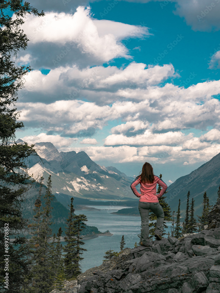 A female hiker stands on the side of a mountain looking at a view of a large reservoir below in Alberta, Canada