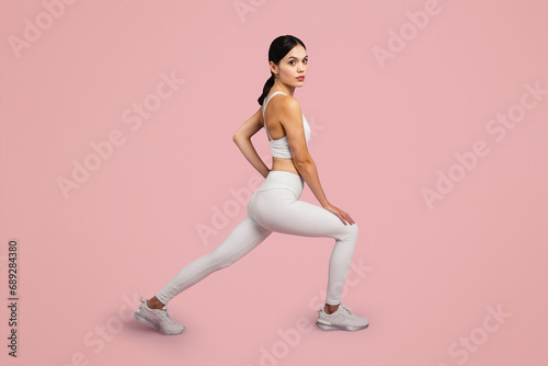 Sporty lady lunging forward exercising during workout over pink studio background, looking at camera, side view
