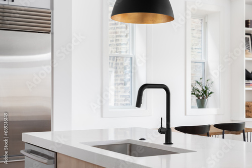 A kitchen sink detail with a black faucet under a black and gold pendant light, white oak island, white countertop, and decorations in the background.	
 photo