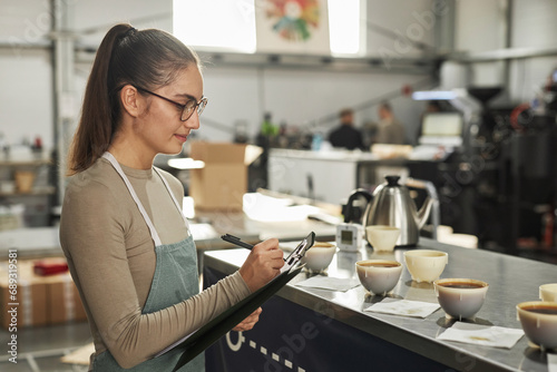 Side view portrait of smiling young woman taking notes on clipboard during cupping and quality control in coffee factory photo