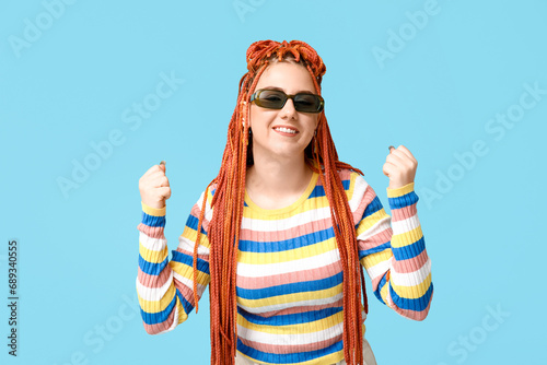 Portrait of fashionable young woman with dreadlocks celebrating success on blue background