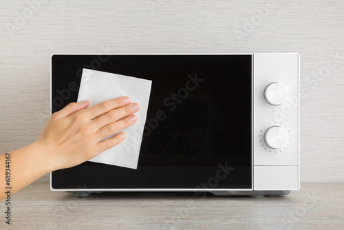Young adult woman hand holding dry white paper napkin and wiping dark black glass door surface of white microwave oven on table top at home kitchen. Closeup. Regular cleanup. Front view. photo