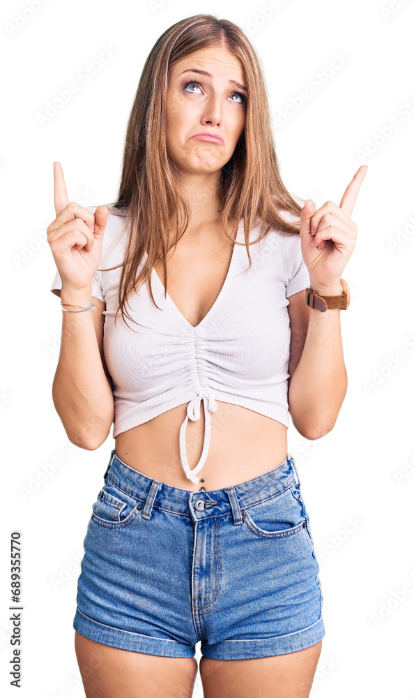 Young beautiful blonde woman wearing casual white tshirt pointing up looking sad and upset, indicating direction with fingers, unhappy and depressed. Young beautiful blonde woman wearing casual white tshirt pointing up looking sad and upset, indicating direction with fingers, unhappy and depressed.