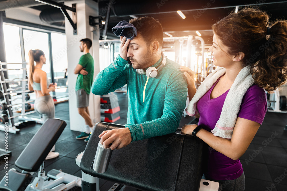 Athletic man taking a break from weight training while his girlfriend is supporting him in the gym. Beautiful woman cheering up her boyfriend during workout. Athletic man taking a break from weight training while his girlfriend is supporting him in the gym. Beautiful woman cheering up her boyfriend during workout.