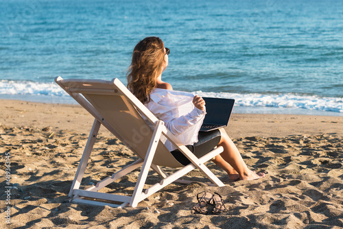 Back view of alone young brunette woman freelancer in office clothes with a laptop looking on the sea sitting on the deck chair on beach. Summer concept © flowertiare