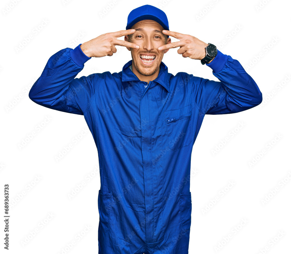 Bald man with beard wearing builder jumpsuit uniform doing peace symbol with fingers over face, smiling cheerful showing victory