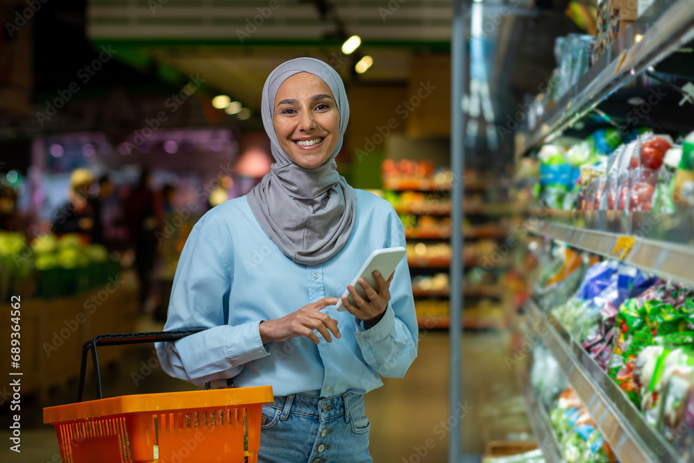 Satisfied Arab woman in hijab is shopping in the store, holding a phone and a shopping basket, smiling and looking at the camera.