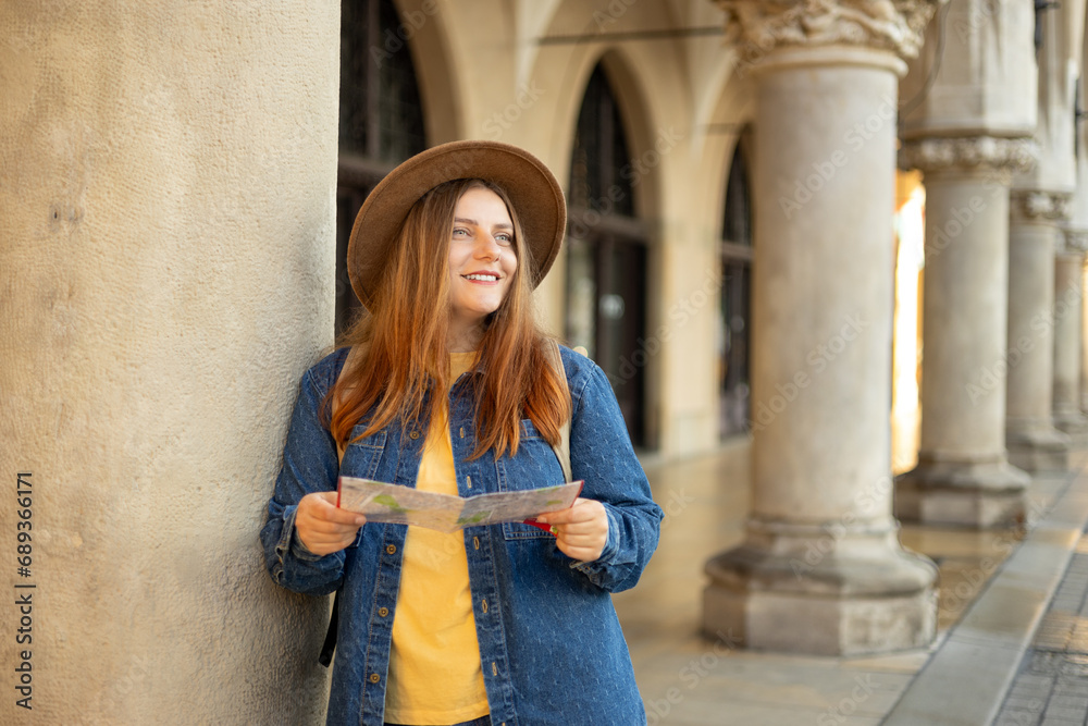 Pretty young woman staying on the street holding paper map in hands. 30s girl in hat posing on Market Square in Krakow. Traveling Europe, High quality photo, The Cloth Hall, Vacation concept
