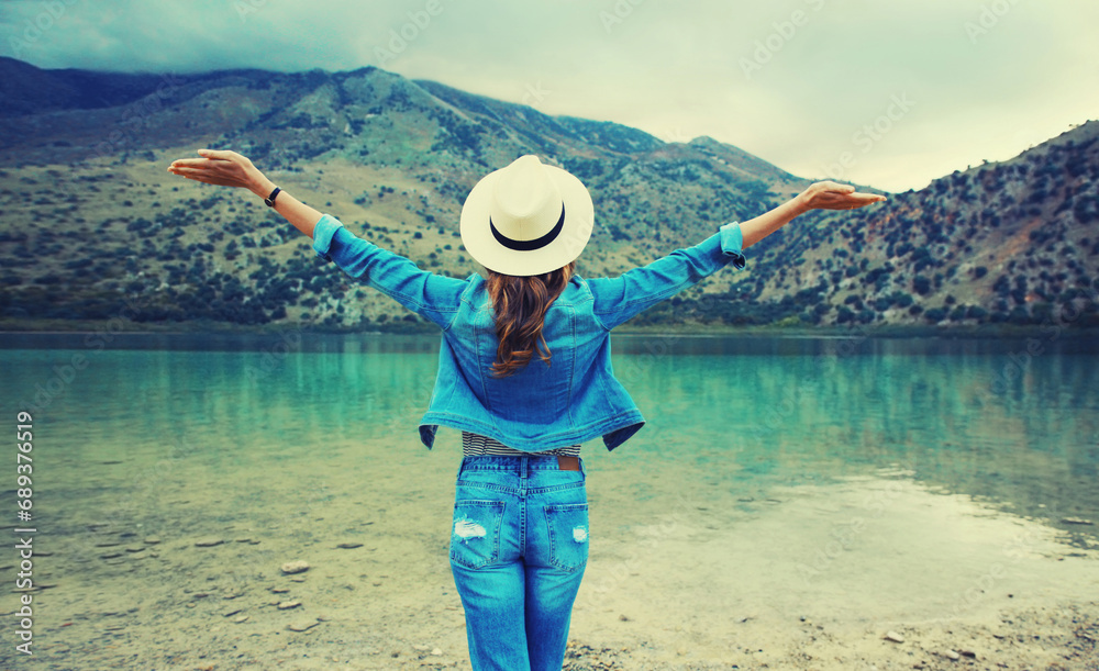 Summer vacation and tourism, rear view of happy woman tourist in straw hat raising her hands up on lake and mountains background. Greece, island Crete.