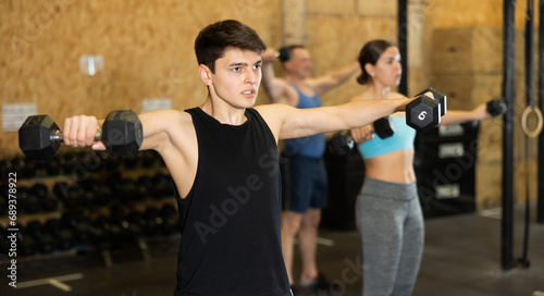 Sportive strong young male in activewear lifting dumbbells during group workout class in gym indoors. Functional training concept