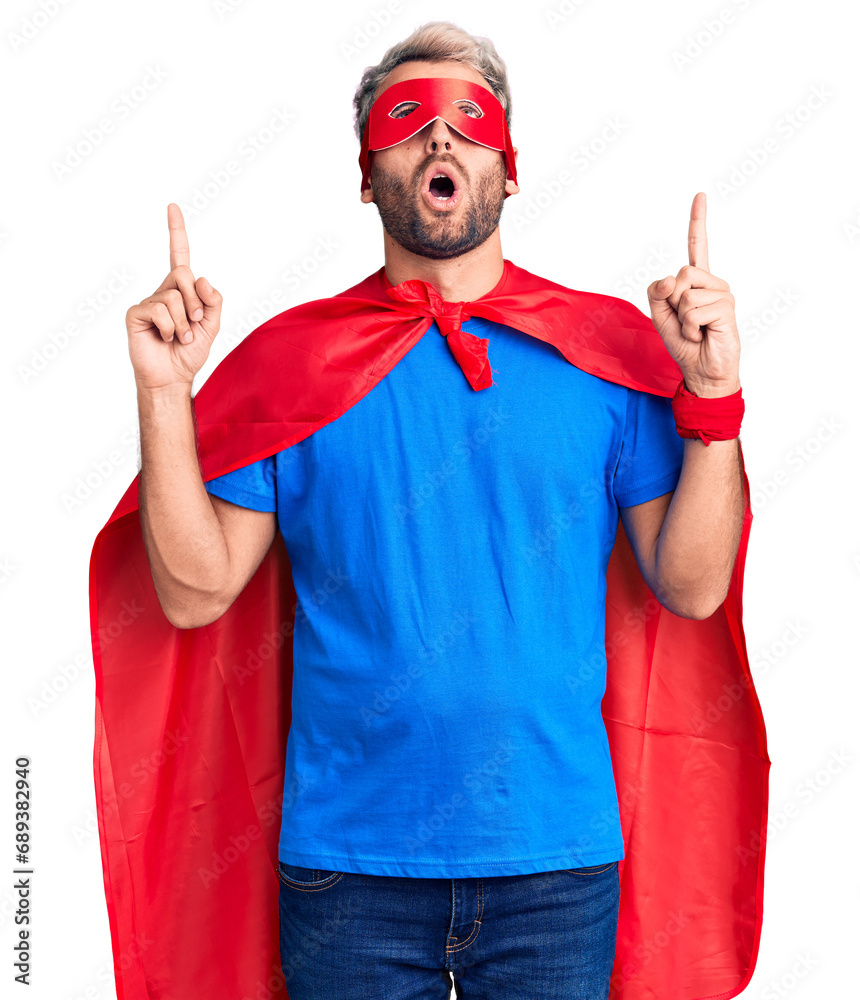 Young blond man wearing super hero custome amazed and surprised looking up and pointing with fingers and raised arms. Young blond man wearing super hero custome amazed and surprised looking up and pointing with fingers and raised arms.