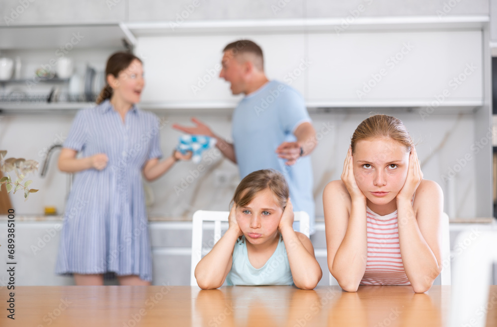 Confused upset daughters sitting at home cover ears with hands during mom and dad quarrel. Children suffer because of parents discord. Husband and wife screaming at each other in background blurred