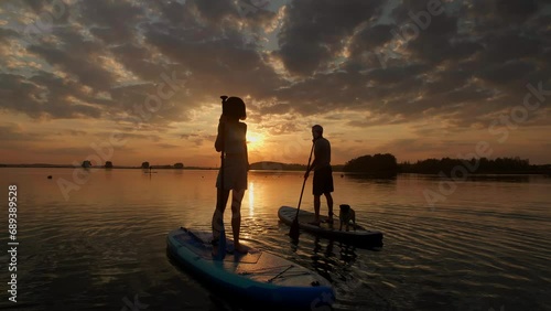 silhouette of couple paddle boarding at lake during sunset together with pug dog. Concept of active family tourism and supping with pets. Brave Dog Standing on SUP Board and enjoy vacation photo