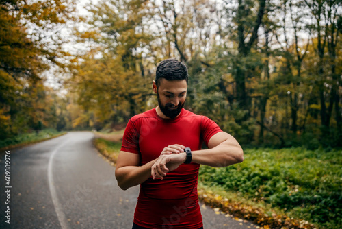 A runner is standing on road in nature and looking at smartwatch.