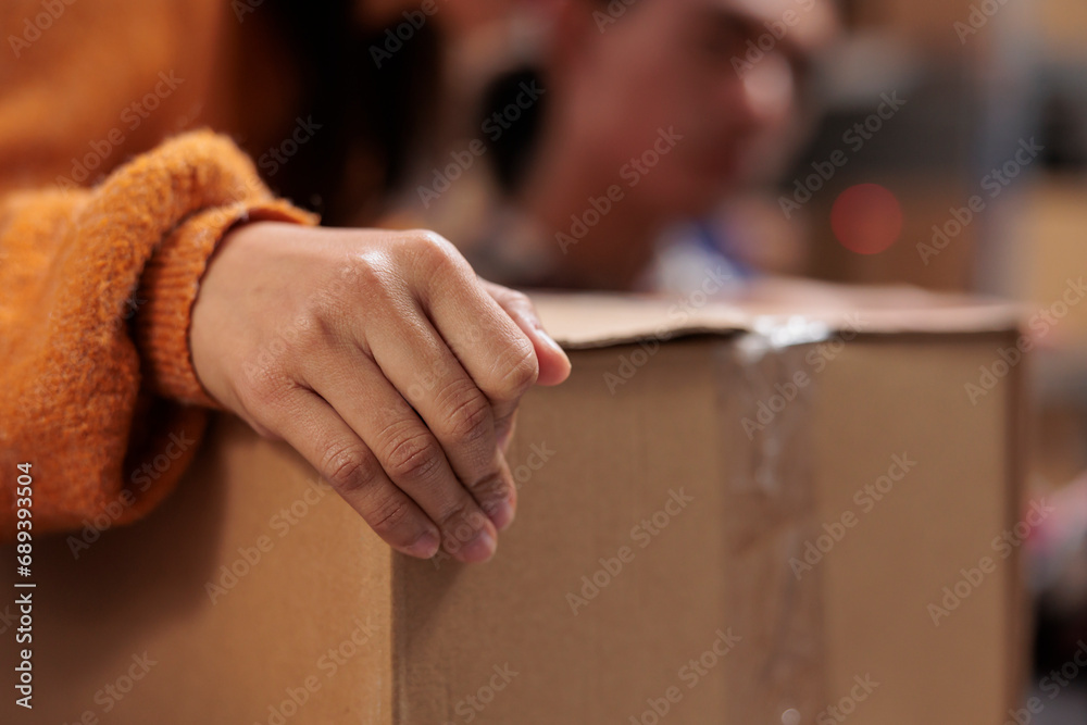 Package handler arm holding packed cardboard box in industrial warehouse. Storehouse employee picking order and preparing customer parcel for transportation with close up on worker hand
