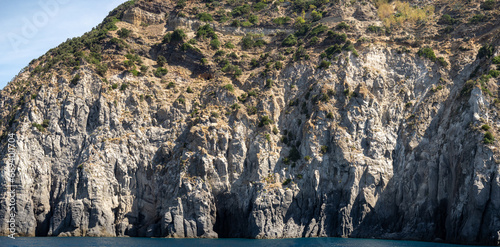 Weathered seaside rock face texture with parts of green and blue water. Aged volcanic stone wall surface background pattern with cracks and scratches. Banner. Ischia Island, Italy.