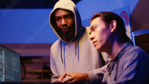 BIPOC hackers in dimly lit room discussing ways to breach defenses and exploit weaknesses in PC systems. Evil cybersecurity gurus in hidden base getting past companies security networks © DC Studio