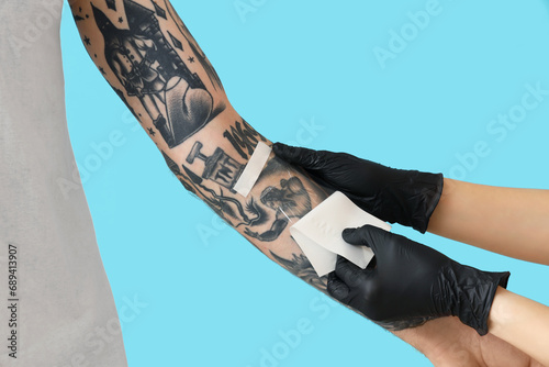Tattoo master in gloves applying protective film cover over fresh tattoo on man's arm against blue background photo