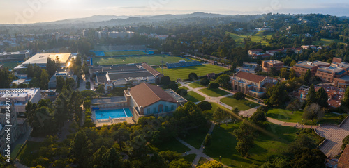 Fotografie Aerial view of UCLA campus bathed in golden light, featuring Gothic architecture, a blue-roofed pool, green hills, and a clear, sunny sky, amidst a vibrant, verdant landscape
