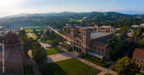 Aerial view of UCLA campus bathed in golden light, showcasing Romanesque Revival and Gothic architecture amid lush greenery, with Royce Hall as the centerpiece.