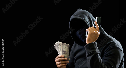 Portrait man thief wearing a black hood shirt, standing hand holding gun and money cash threaten, counting the amount obtained from hijacking or robbing, in pitch-black room. dark background photo