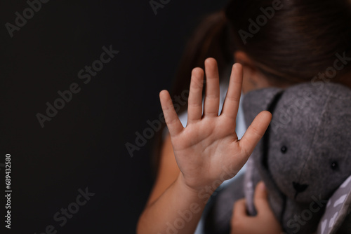 Child abuse. Little girl with toy bunny doing stop gesture on dark background, selective focus. Space for text photo