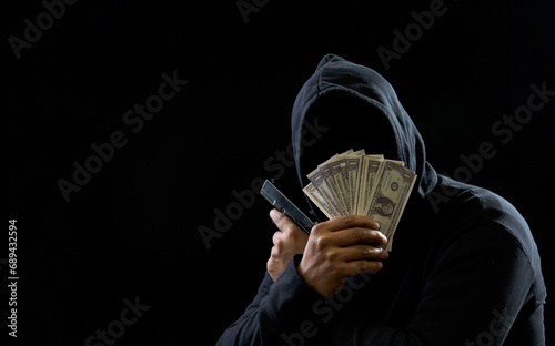 Portrait man thief wearing a black hood shirt, standing hand holding gun and money cash threaten, counting the amount obtained from hijacking or robbing, in pitch-black room. dark background photo