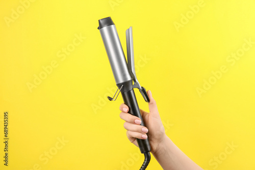 Hair styling appliance. Woman holding curling iron on yellow background, closeup