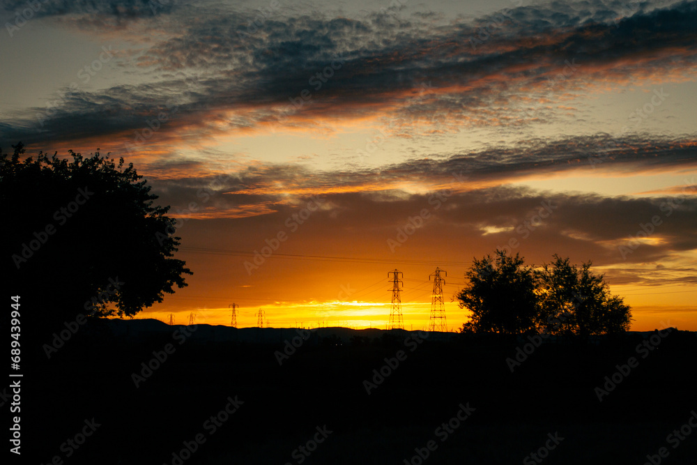 Fototapeta premium Orange nature sunset with huge power poles on horizon with silhouette of trees and evening clouds in California landscape in Usa, America