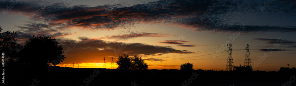 Fototapeta premium Orange nature sunset with huge power poles on horizon with silhouette of trees and evening clouds in California landscape in Usa, America