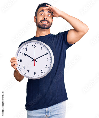 Young hispanic man holding big clock stressed and frustrated with hand on head, surprised and angry face