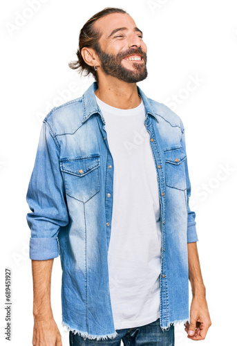 Attractive man with long hair and beard wearing casual denim jacket looking away to side with smile on face, natural expression. laughing confident.