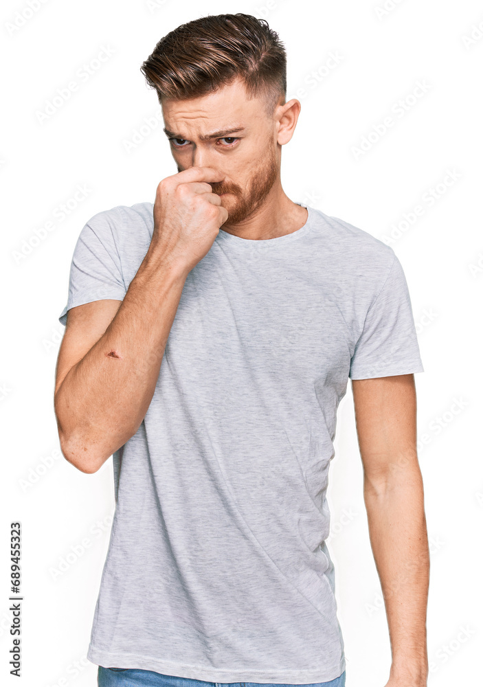 Young redhead man wearing casual grey t shirt smelling something stinky and disgusting, intolerable smell, holding breath with fingers on nose. bad smell Young redhead man wearing casual grey t shirt smelling something stinky and disgusting, intolerable smell, holding breath with fingers on nose. bad smell