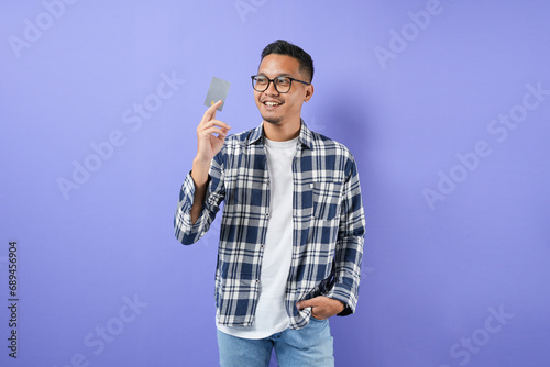 Portrait of a smiling Asian man with a credit card in his hand. Non-cash payment concept and isolated on a purple background. 