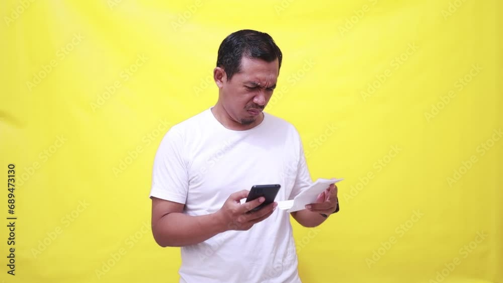 Confused asian man standing while comparing shopping receipt with his phone. Isolated on yellow background Confused asian man standing while comparing shopping receipt with his phone. Isolated on yellow background