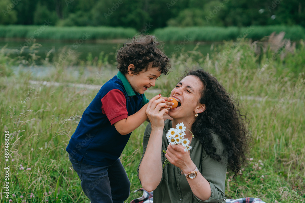 Fototapeta premium Cheerful little boy having fun with mom feeds her by fruits laughing standing on lawn against river and forest on background. Family leisure, vacations together.