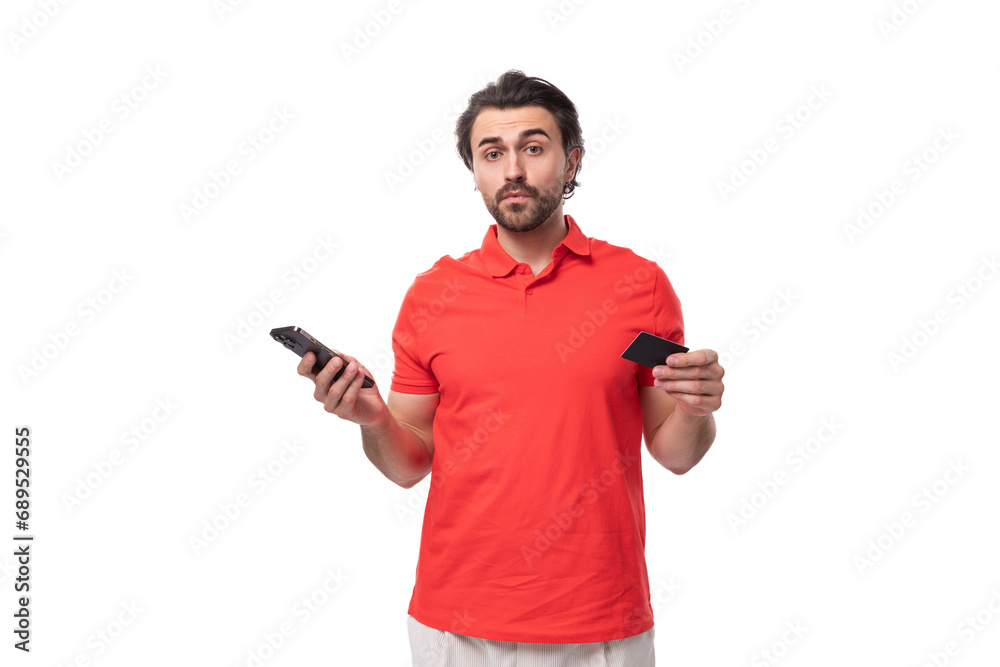 a young brutal caucasian brunette man with a beard dressed in red clothes uses a smartphone and a plastic bank card a young brutal caucasian brunette man with a beard dressed in red clothes uses a smartphone and a plastic bank card