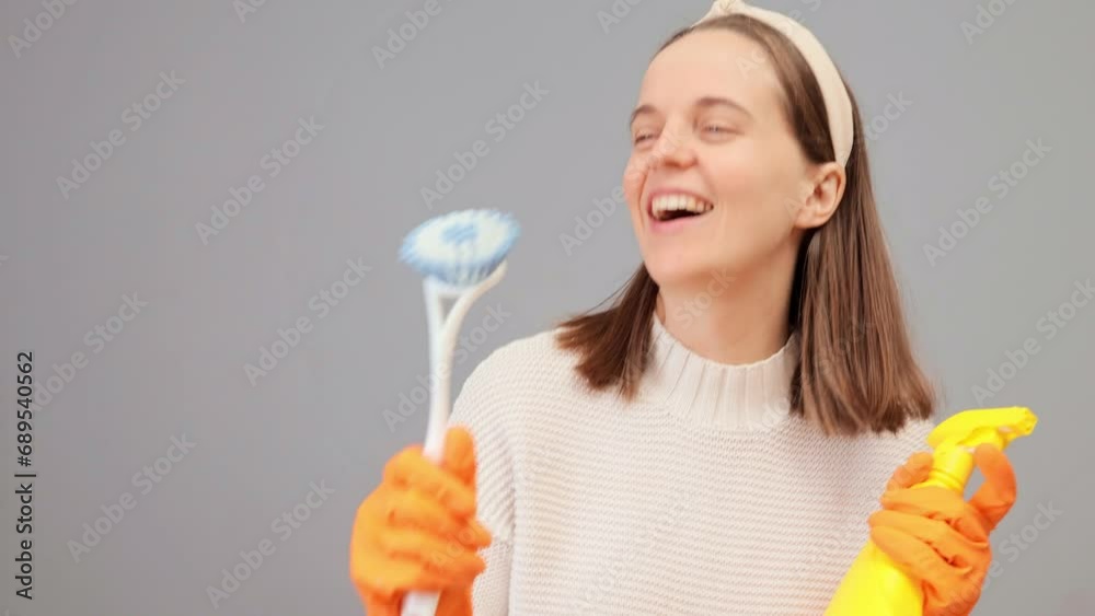 Happy woman in jumper hair band and protective gloves holding brush and detergent imagining she is star singing and dancing while doing her housekeeping routine posing isolated over gray background Happy woman in jumper hair band and protective gloves holding brush and detergent imagining she is star singing and dancing while doing her housekeeping routine posing isolated over gray background