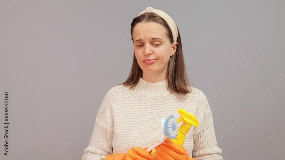 Despair woman in orange rubber gloves dislike to clean house looking around with brush and detergent bottle thinking where to begin her housework standing isolated over gray background Despair woman in orange rubber gloves dislike to clean house looking around with brush and detergent bottle thinking where to begin her housework standing isolated over gray background