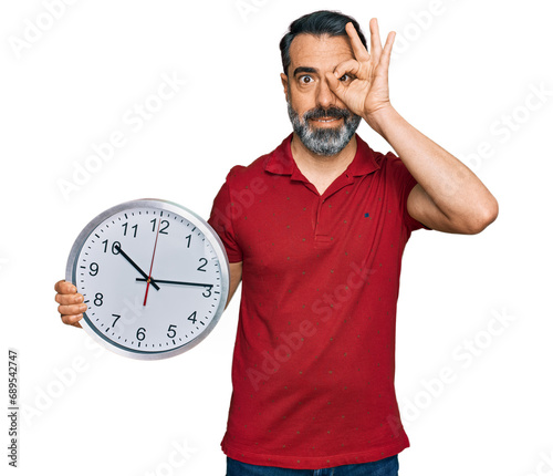 Middle aged man with beard holding big clock smiling happy doing ok sign with hand on eye looking through fingers