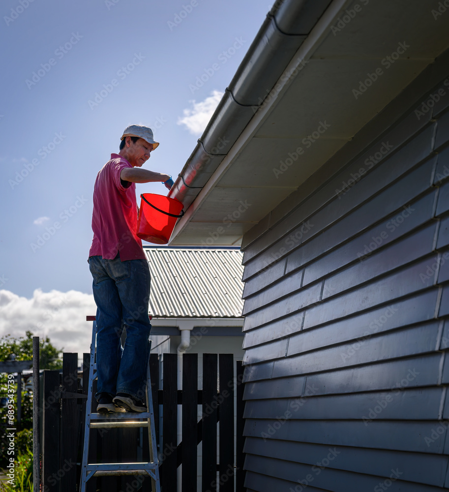 Man standing on the ladder and cleaning the gutter. Home maintenance work. Vertical format. Auckland.