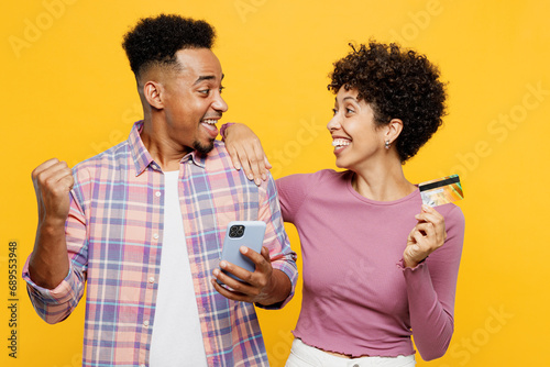 Young couple two friends family man woman wear purple casual clothes together use mobile cell phone hold credit bank card shopping online do winner gesture isolated on plain yellow orange background.