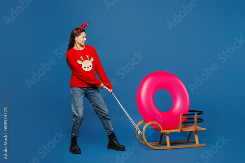Traveler woman wear red Christmas sweater carry rubber ring on sled isolated on plain blue background. Tourist travel abroad in free time rest getaway. Air flight trip Happy New Year holiday concept. #689554778