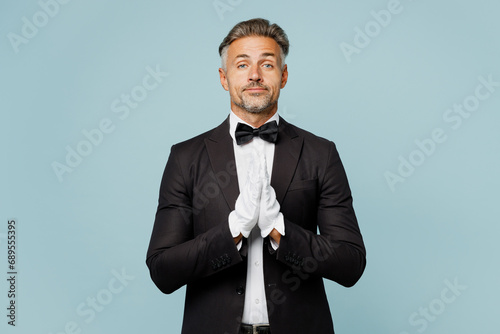 Adult barista male waiter butler man wear shirt black suit bow tie elegant uniform hold hands folded in prayer gesture beg work at cafe isolated on plain blue background. Restaurant employee concept. photo