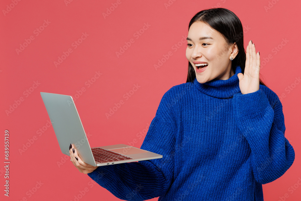 Young IT woman of Asian ethnicity wears blue sweater casual clothes hold use work on laptop pc computer waving hand isolated on plain pastel light pink background studio portrait. Lifestyle concept. Young IT woman of Asian ethnicity wears blue sweater casual clothes hold use work on laptop pc computer waving hand isolated on plain pastel light pink background studio portrait. Lifestyle concept.