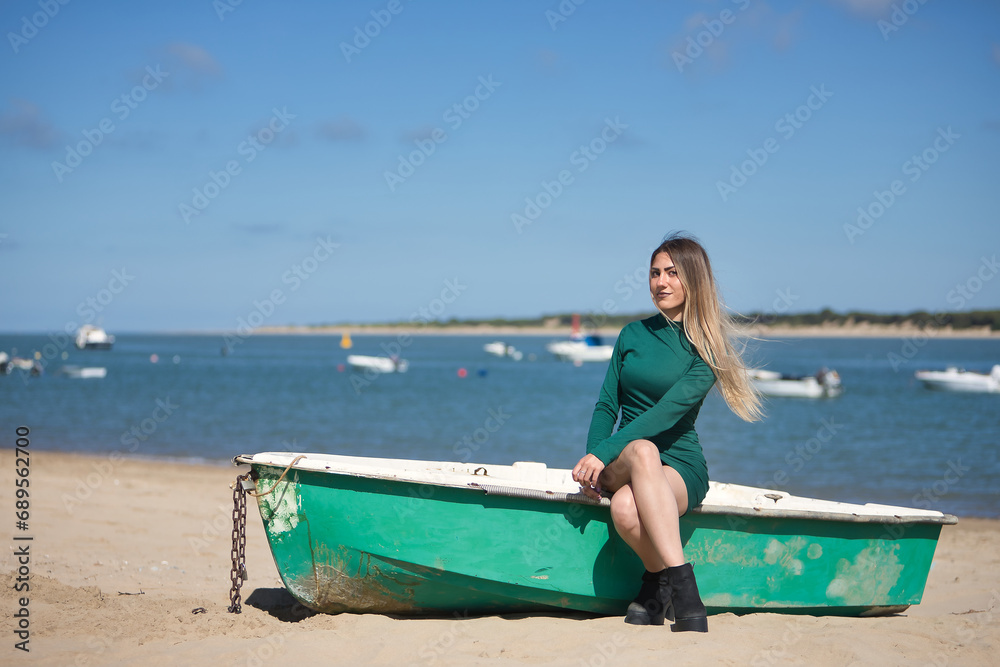 Fototapeta premium Young, pretty, blonde, green-eyed young woman in a green dress, sitting in a boat, looking at the camera, relaxed and calm, on the beach, with the sea in the background. Concept vacation, sun, travel