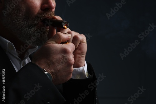 Bearded man lighting cigar on dark grey background, closeup. Space for text