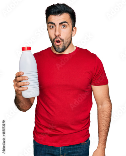 Hispanic man with beard holding liter bottle of milk scared and amazed with open mouth for surprise, disbelief face