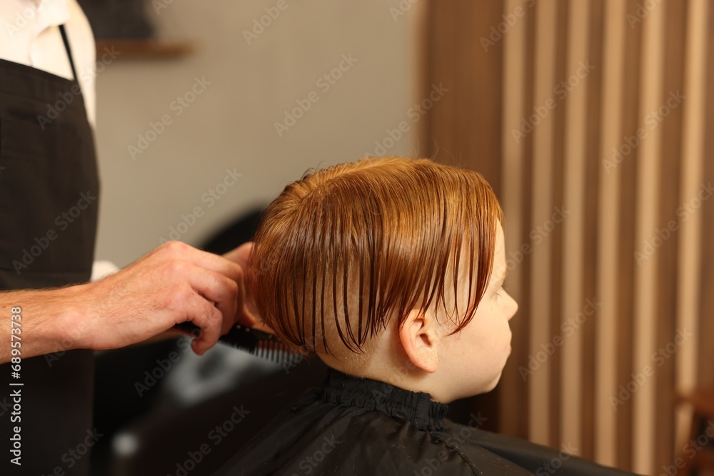 Naklejka premium Professional hairdresser combing boy's hair in beauty salon, closeup